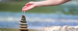 Рука человека аккуратно удерживает капли воды над аккуратно сложенной пирамидой из камней на берегу водоема.
