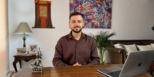 Мужчина в коричневой рубашке сидит за деревянным столом с ноутбуком, на фоне уютного интерьера с растением, картинами и лампой.