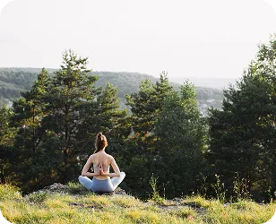Женщина занимается йогой на природе, сидя на скале с видом на лесистый пейзаж и горы.